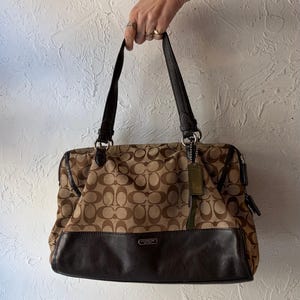 以下が含まれることがあります： 茶色とタン色のコーチハンドバッグ。ロゴパターンが繰り返されています。バッグは濃い茶色の革のベースとハンドルが付いています。グリーンのタグがハンドルからぶら下がっています。