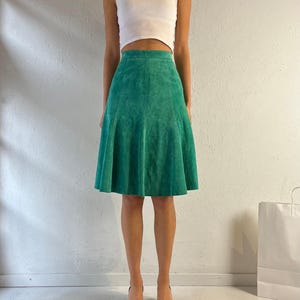 Può includere: Una gonna di camoscio verde con un orlo svasato. La gonna è lunga fino al ginocchio e ha un'alta vita.