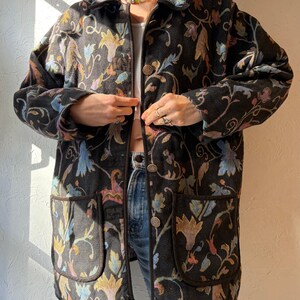 May include: Black jacket with a floral pattern in yellow, blue, and pink. The jacket has two front pockets and brown buttons. The jacket is a button-down style.