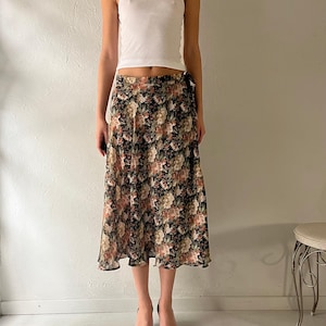 Peut inclure: Un débardeur blanc associé à une jupe midi à imprimé floral. La jupe présente un fond noir avec un motif de fleurs roses, pêche et vertes. Des talons noirs à bout pointu avec des détails de boucle complètent la tenue.