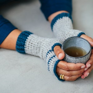May include: A pair of gray and teal fingerless gloves with a crocheted design. The gloves are worn by a person holding a gray ceramic mug with a warm beverage inside.