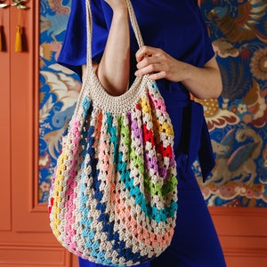May include: A colorful, handmade crochet tote bag. The bag features a beige base with vibrant stripes of red, blue, green, yellow, and pink. The bag is held by a person wearing a blue outfit.