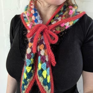 Puede incluir: Bufanda de ganchillo hecha a mano con un diseño de cuadrados de la abuela. La bufanda presenta una vibrante mezcla de colores, incluyendo azul, amarillo, rosa y verde, delineados en rojo. La bufanda está atada con un cordón rojo.