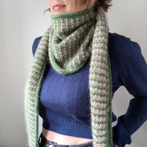 May include: A green and pink knitted scarf with a geometric pattern. The scarf is wrapped around the neck and drapes down the front. The scarf is made of a soft, fuzzy material. The background is a light grey colour.