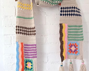 May include: A beige crocheted scarf with colorful geometric patterns and tassels. The scarf features stripes, squares, and polka dots in shades of pink, blue, green, yellow, orange, brown, and purple.