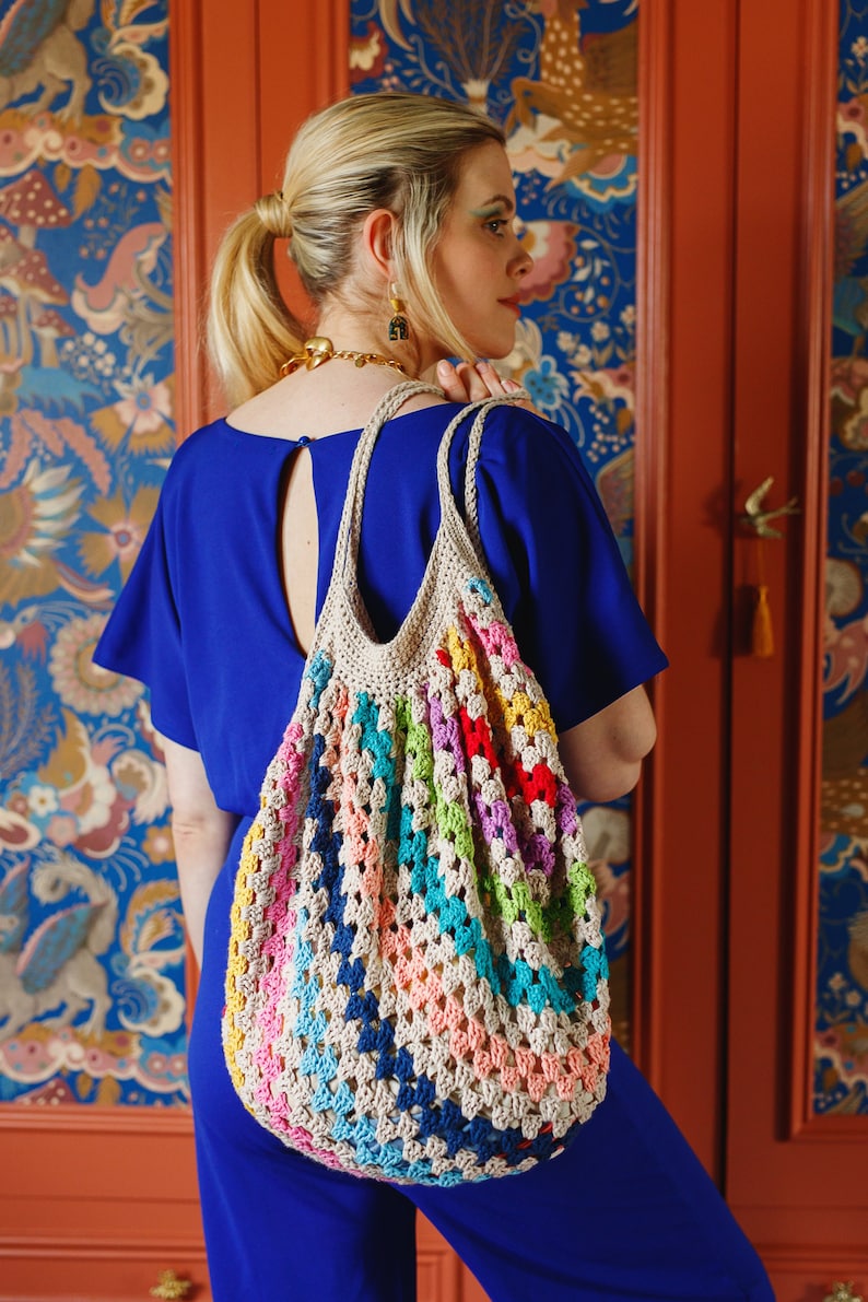 May include: A colorful crochet tote bag with a beige handle and a vibrant pattern of rainbow colors. The bag is carried over the shoulder, showcasing its handmade design and open weave. The bag's dimensions are approximately 15 inches wide and 18 inches tall.
