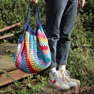 May include: A colorful, handmade crochet tote bag with a blue handle. The bag features a striped pattern in various colors, including red, yellow, purple, and blue. The bag is held by a person wearing gray jeans and sneakers.