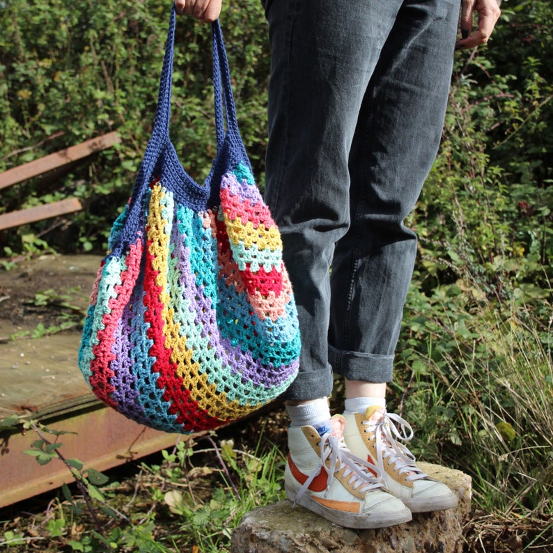 May include: A colorful, handmade crochet tote bag with a blue handle. The bag features a striped pattern in various colors, including red, yellow, purple, and blue. The bag is held by a person wearing gray jeans and sneakers.