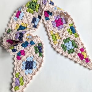 Może przedstawiać: Ręcznie robiony szalik z szydełka w kształcie trójkąta. Szalik ma jasnobeżową podstawę z kolorowymi kwadratowymi wzorami w odcieniach różu, niebieskiego, zieleni i żółci. Krawędzie mają falisty wzór.