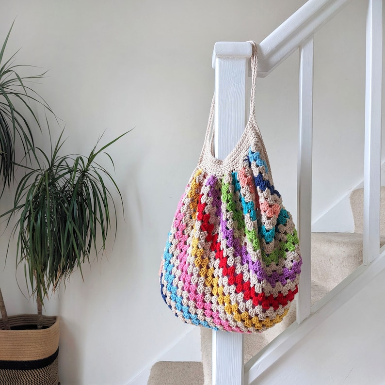 May include: A colorful, handmade crochet tote bag. The bag features a granny square design with a variety of colors including red, blue, green, and yellow. The bag has a beige handle and trim, and is hanging on a white banister.