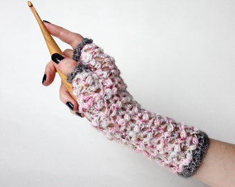 May include: Pink and white crocheted fingerless gloves with a gray trim. A wooden crochet hook is held in the hand wearing the gloves.