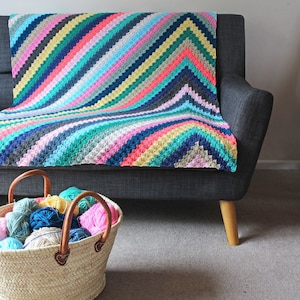 Puede incluir: Una manta de crochet colorida con un patrón de chevron en tonos de rosa, azul, verde y amarillo. La manta está drapeada sobre un sofá gris y una cesta de hilo está en primer plano.