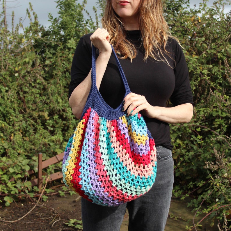 May include: A colorful, handmade crochet tote bag. The bag features a rainbow of colors including red, yellow, blue, and purple. The bag has a navy blue rim and straps. The bag is held by a person.