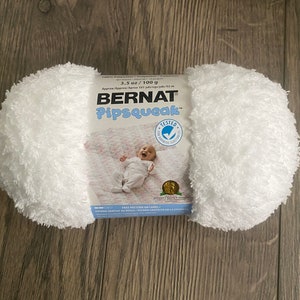 May include: A skein of white Bernat Pipsqueak yarn, a soft, fluffy yarn for crocheting or knitting baby blankets and other projects. The yarn label features a baby in a white onesie and the text "Bernat Pipsqueak" and "Tested for harmful substances".