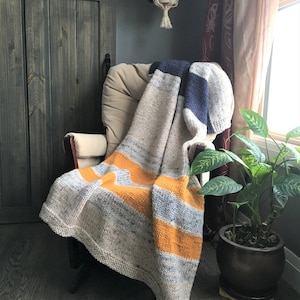 May include: A knitted blanket with beige, orange, and blue stripes draped over a chair. The chair is beige, and the background includes a wooden door, a plant, and a window with curtains. The blanket is likely made of wool or a similar yarn.