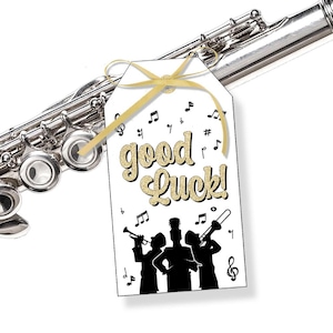 May include: A silver flute with a white tag tied with a gold ribbon. The tag has the words "good Luck!" in gold glitter lettering, musical notes, and a silhouette of a band playing instruments.