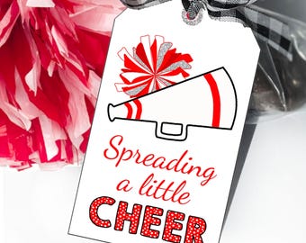 May include: A white rectangular gift tag featuring a red and silver glitter cheerleader pom-pom illustration above a red-lined megaphone graphic. The text reads Spreading a little CHEER in red script and bold red polka-dot lettering with a buffalo plaid ribbon.