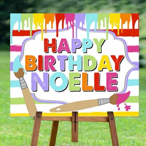 May include: A colourful birthday sign with paint dripping down the sides. The sign says "Happy Birthday Noelle" in rainbow letters. Two paintbrushes are on either side of the sign.