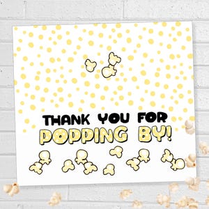 May include: A white card with a yellow popcorn and polka dot design. The text "THANK YOU FOR POPPING BY!" is displayed in black and yellow. Several popcorn kernels are scattered around the card.