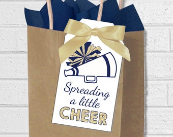 Puede incluir: Una bolsa de regalo de papel marrón con papel de seda azul marino y una cinta dorada. Una etiqueta blanca con un diseño de pompón y megáfono azul marino y dorado está adjunta. La etiqueta dice "Spreading a little CHEER."