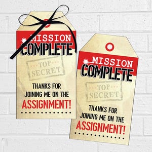 May include: Two rectangular paper gift tags with a cream-coloured background and red top border. The text reads MISSION COMPLETE, TOP SECRET, and THANKS FOR JOINING ME ON THE ASSIGNMENT! A black ribbon is tied through the hole of the left tag.