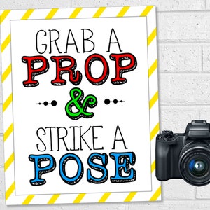 May include: A white sign with yellow stripes on the border. The sign says "Grab a Prop & Strike a Pose" in colorful letters. A black camera is to the right of the sign.