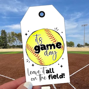 Può includere: Un'etichetta regalo rettangolare bianca con un foro nella parte superiore, caratterizzata da una grafica di una palla da softball gialla con cuciture rosse. Il testo recita "it's game day" in nero, seguito da "Leave it all on the field!" in nero.
