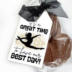 May include: A rectangular white gift tag with a black ribbon bow, featuring a black silhouette of a gymnast in a leap pose over gold glitter, with the text "it's a GREAT TIME To have our BEST DAY!" attached to a clear bag of chocolate treats.