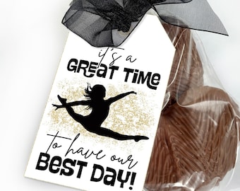May include: A rectangular white gift tag with a black ribbon bow, featuring a black silhouette of a gymnast in a leap pose over gold glitter, with the text "it's a GREAT TIME To have our BEST DAY!" attached to a clear bag of chocolate treats.