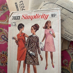 May include: Vintage Simplicity sewing pattern, number 7033, featuring illustrations of three women in dresses. The pattern includes text such as "Jr. Petite" and "Bust 32 1/2". The background is a pink paisley fabric.
