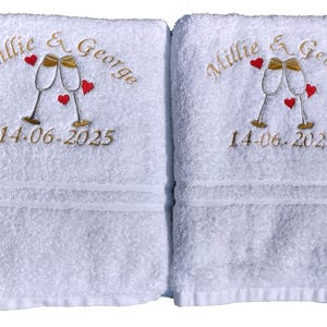 May include: Two white bath towels with embroidered gold text and design. The design includes two champagne flutes, red hearts, and the names "Millie & George" with the date "14.06.2025".