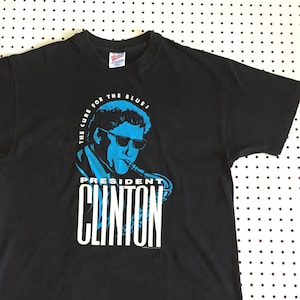 May include: Black t-shirt with a blue graphic of President Clinton playing the saxophone. The graphic says "The Cure for the Blues" and "President Clinton".