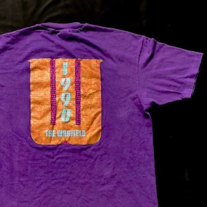 Puede incluir: Camiseta morada con un gráfico de una gran letra naranja "U" con el texto "1999" y "The Warfield" impreso en ella.