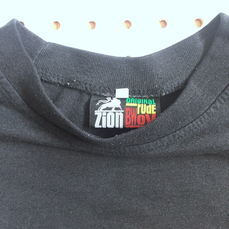 以下が含まれることがあります： 黒いTシャツに、黒、緑、黄、赤のラベルが付いており、「Original Rudebwoy Zion」と書かれています。