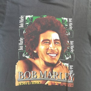 以下が含まれることがあります： ボブ・マーリーのカラフルな画像がプリントされた黒いTシャツ。画像には「ボブ・マーリー」と「ナチュラル・ミスティック」の文字が含まれています。