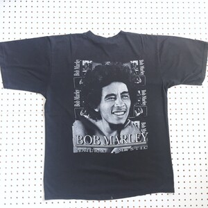 以下が含まれることがあります： ボブ・マーリーの白黒画像がプリントされた黒いTシャツ。画像は「ボブ・マーリー」というテキストで囲まれており、「ナチュラル・ミスティック」という文字が画像の下に印刷されています。