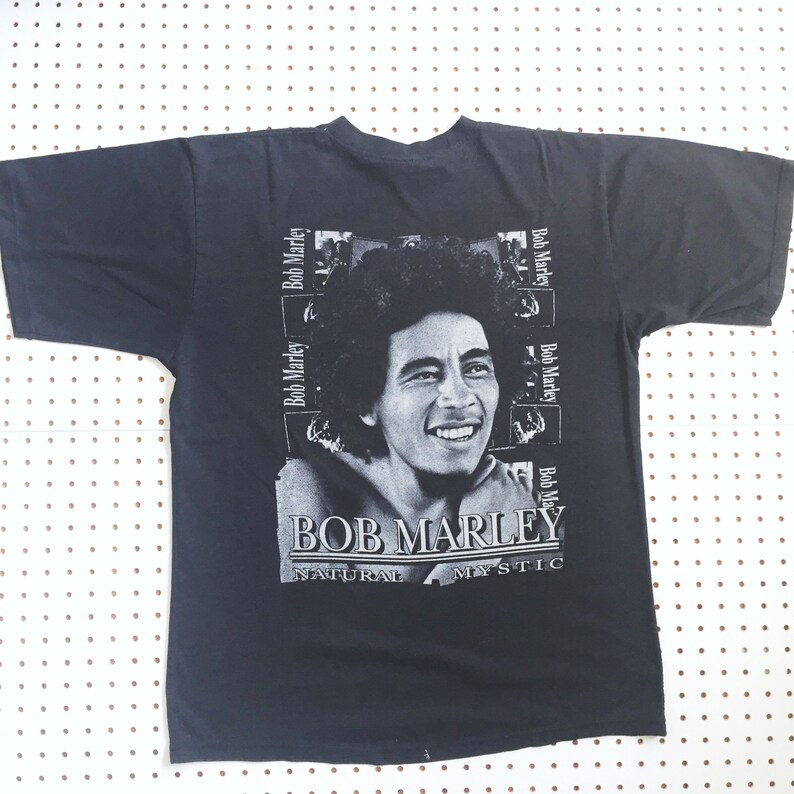 以下が含まれることがあります： ボブ・マーリーの白黒画像がプリントされた黒いTシャツ。画像は「ボブ・マーリー」というテキストで囲まれており、「ナチュラル・ミスティック」という文字が画像の下に印刷されています。