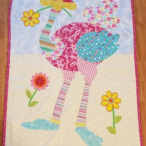 Könnte beinhalten: Eine bunte Patchwork-Decke mit einer rosa und blauen Strauß mit gelbem Schnabel und blauen Augen. Der Strauß ist von gelben, roten und grünen Blumen umgeben. Die Decke hat einen weißen Hintergrund mit rosa Punkten.