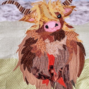 Può includere: Una mucca Highland marrone e bianca con il naso rosa e gli occhi neri, realizzata con ritagli di stoffa, è in piedi su uno sfondo verde e bianco.