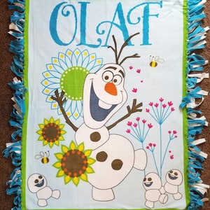 May include: A blue and green tie-dye blanket with a white background featuring Olaf from Frozen. The blanket has a green border and blue fringe. The image shows Olaf with a happy expression, surrounded by sunflowers and bees. The text "OLAF" is written in blue at the top of the blanket.