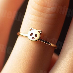 May include: A gold-colored ring featuring a panda bear design. The panda's face is detailed with black and white enamel. The ring is delicate and sits on a finger.
