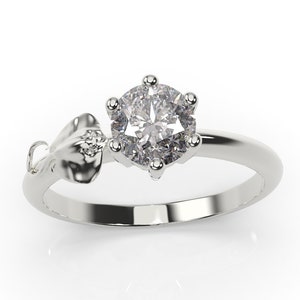 May include: A silver ring with a large, round, clear gemstone set in a six-prong setting. The ring has a delicate, floral design on the side.