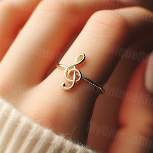 May include: A gold-tone ring with a treble clef design. The ring is worn on a finger.
