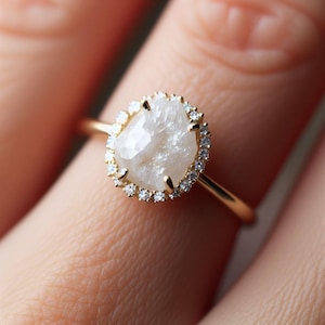 Op de afbeelding: Een gouden ring met een grote, onregelmatig gevormde, witte edelsteen, omringd door kleine, heldere edelstenen. De ring heeft een dunne gouden band. De ring zit om een vinger.