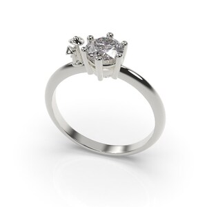 May include: A silver ring with a large, clear, round gemstone set in a prong setting. There are smaller gemstones on the side of the ring.