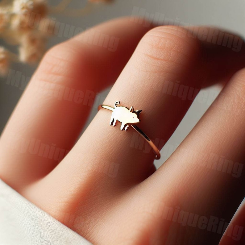 May include: A gold-toned ring with a small pig charm. The ring is on a person's finger.
