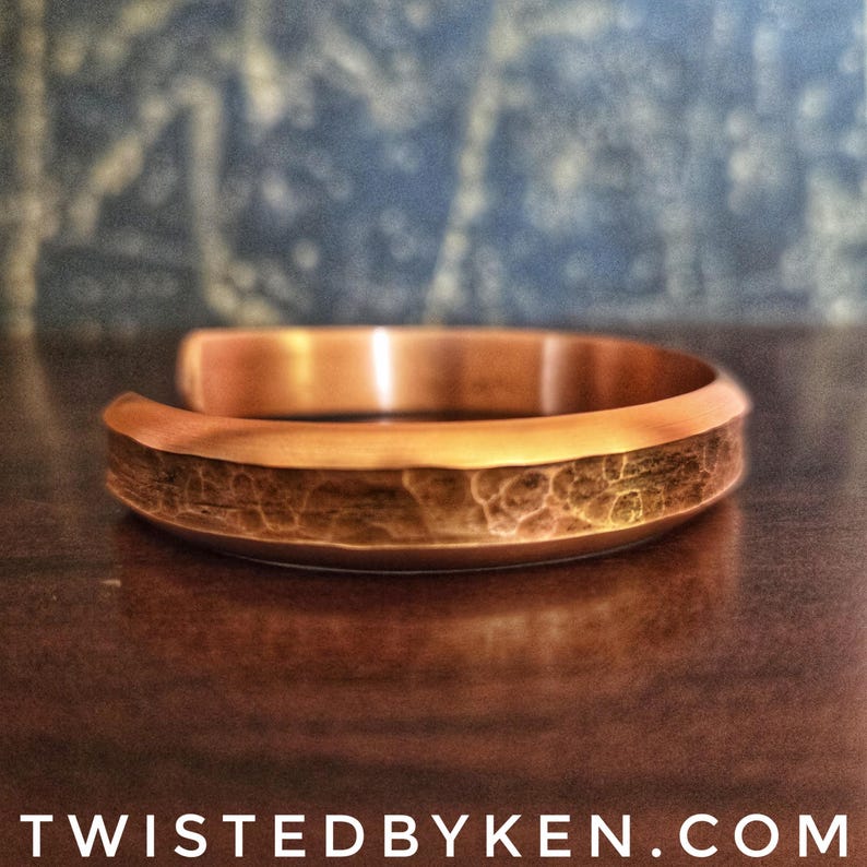 Puede incluir: Un brazalete de cobre martillado con una forma redondeada ligeramente curvada. El brazalete tiene un acabado martillado texturizado.