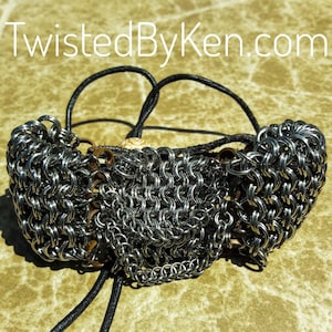 May include: A silver chainmail bracelet with a brown leather cord and a white bead. The bracelet is made of multiple chainmail patterns and is tied together with the leather cord. The text "TwistedByKen.com" is visible in the background.