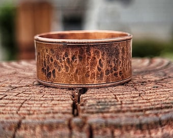 May include: A copper ring with a hammered texture and a decorative border. The ring is sitting on a wooden surface.