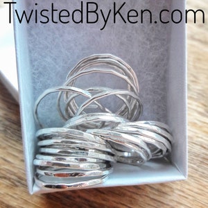 May include: A collection of silver rings with a hammered texture, displayed in a white box. The rings are stacked and intertwined, showcasing a unique, handcrafted design. The website address "TwistedByKen.com" is visible in the top left corner.